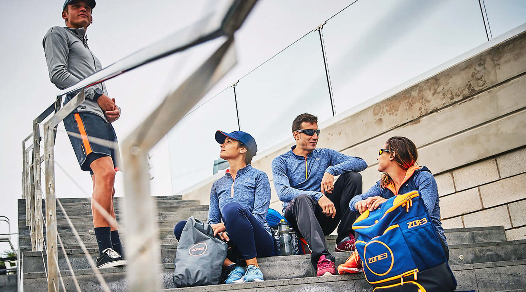 Group of Zone3 athletes in blue and gray training wear sits on stairs with bags after a workout.