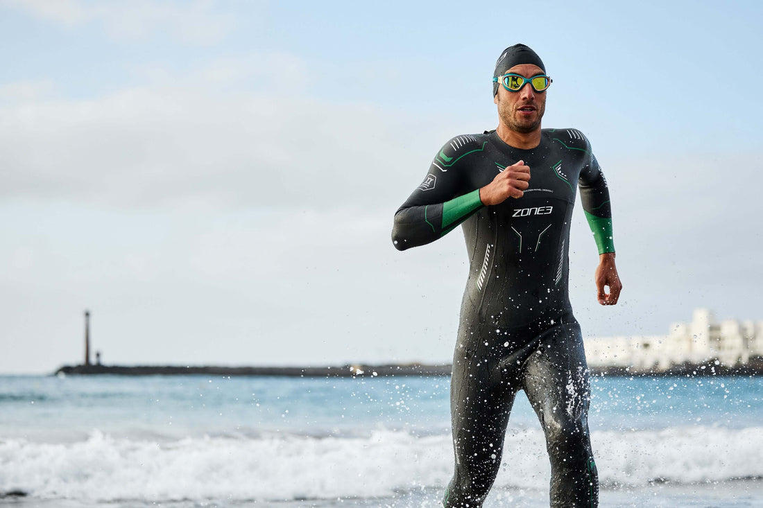 Male athlete in a black and green tri suit, wearing a swim cap and mirrored goggles, runs along the beach with water spray.