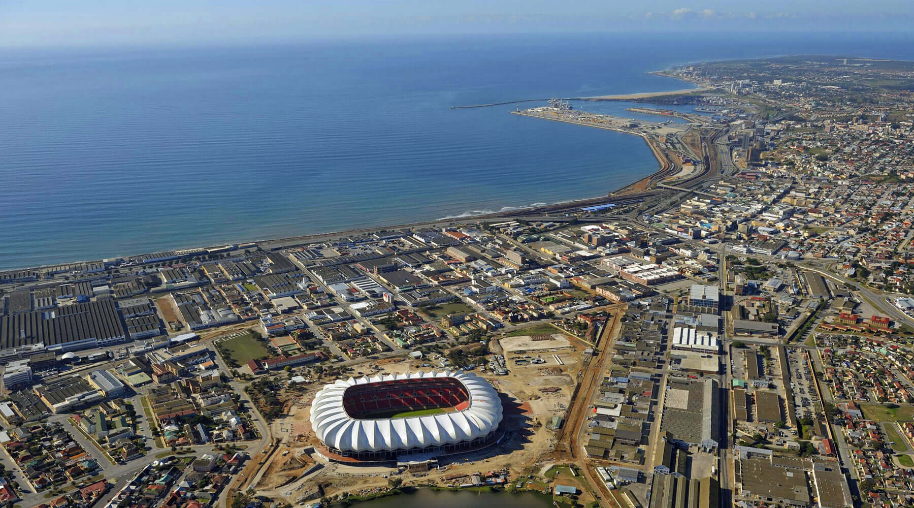 Aerial view of a coastal city with a large stadium in the foreground and dense urban development along the shoreline.