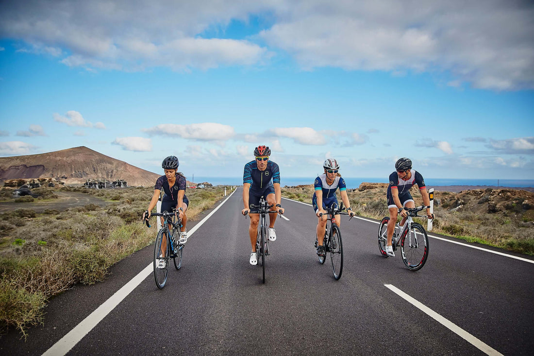 Cyclists ride together on a coastal highway under blue skies with hills in the distance