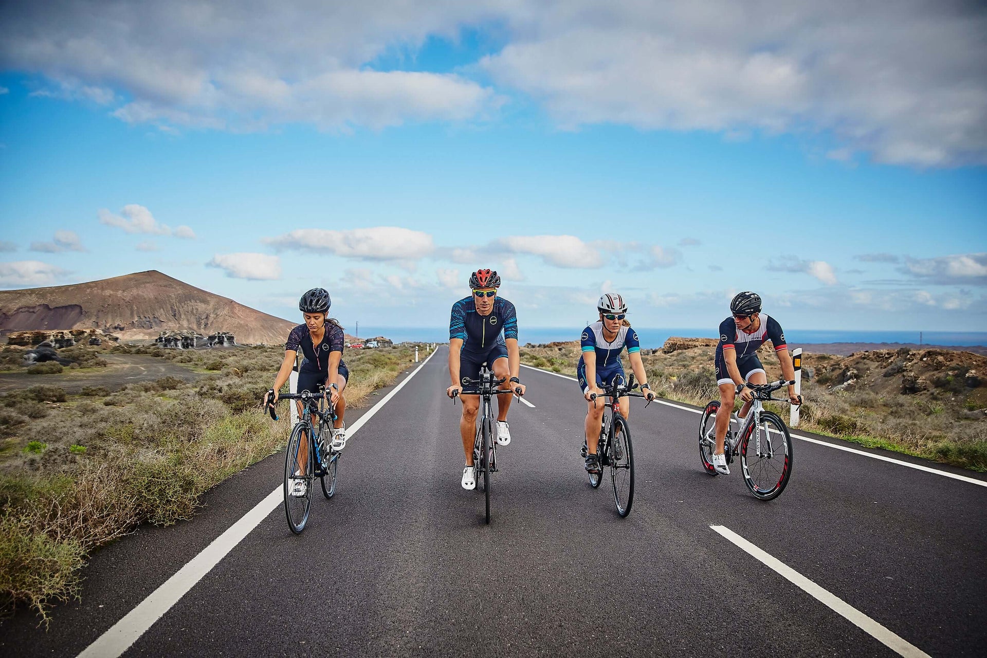 Cyclists ride together on a coastal highway under blue skies with hills in the distance