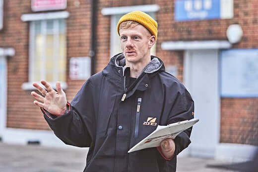 Man in a dark jacket and orange beanie gestures while holding a clipboard on an urban street.