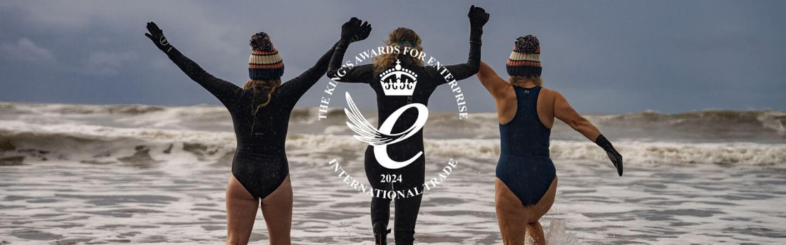 Three swimmers in wetsuits celebrate on a windy beach with a circular award logo centered.