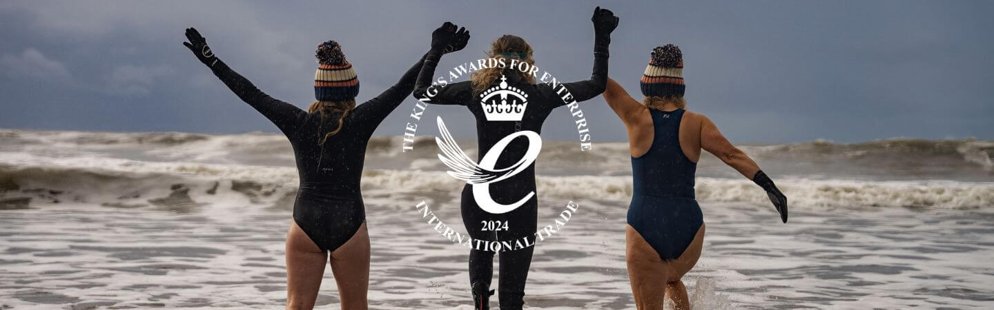Three swimmers in wetsuits celebrate on a windy beach with a circular award logo centered.