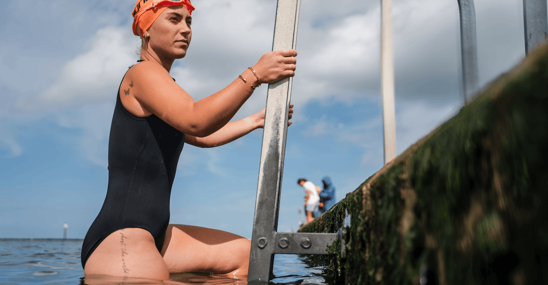 Female swimmer in a black wetsuit and orange swim cap grips a metal ladder at the water’s edge before entering.