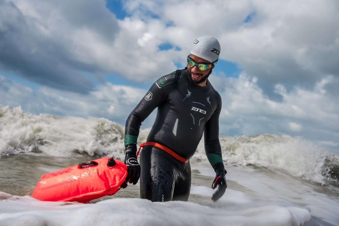 Open water swimmer in a white cap and black wetsuit rides a wave while holding a bright red safety buoy.