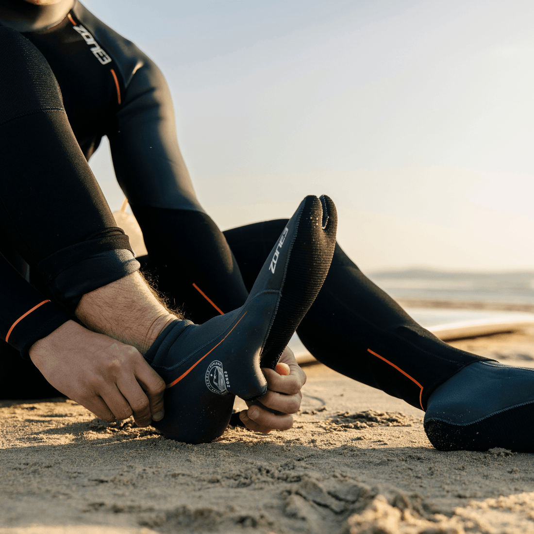 A swimmer sitting on a beach pulling on black ZONE3 neoprene swim socks.