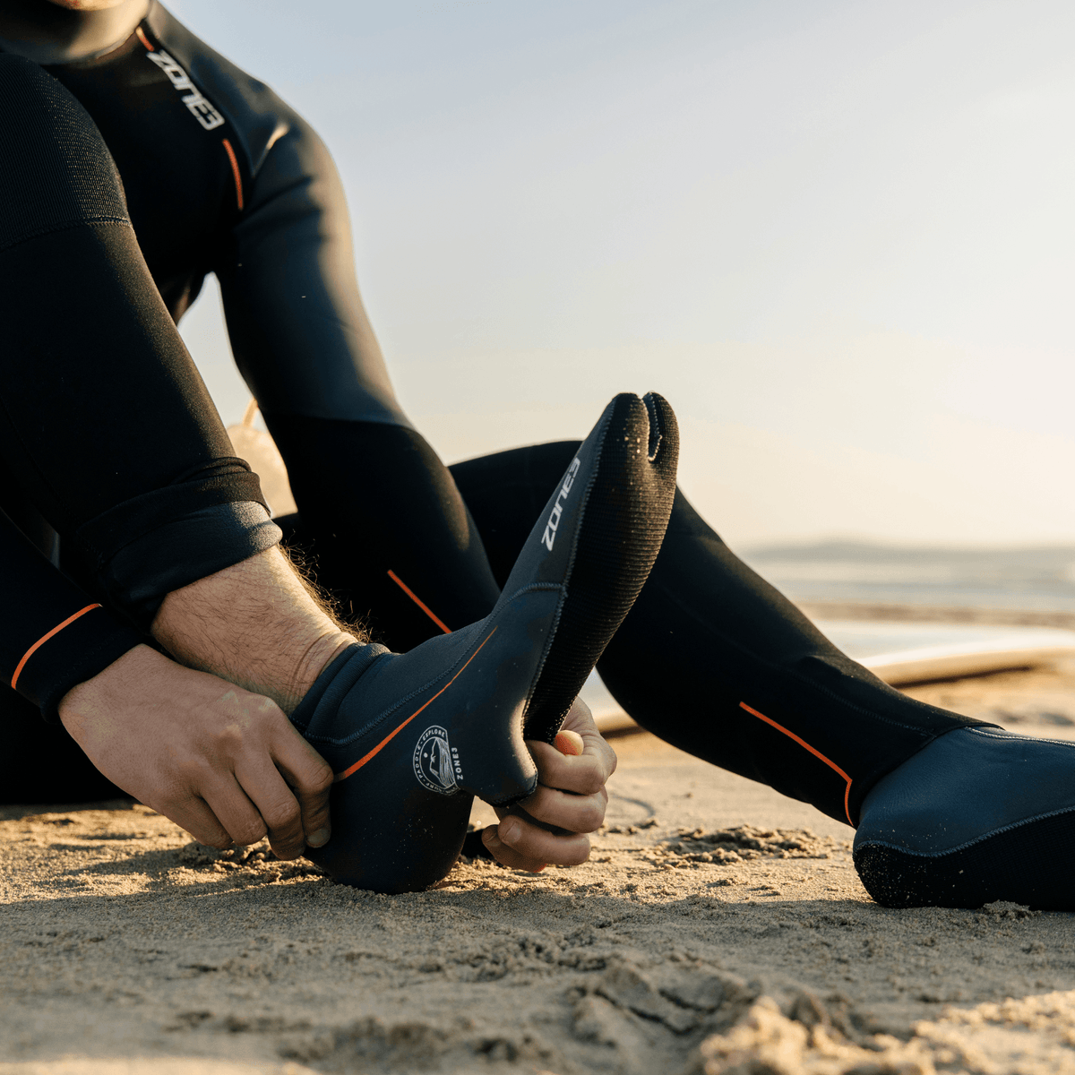 A swimmer sitting on a beach pulling on black ZONE3 neoprene swim socks.