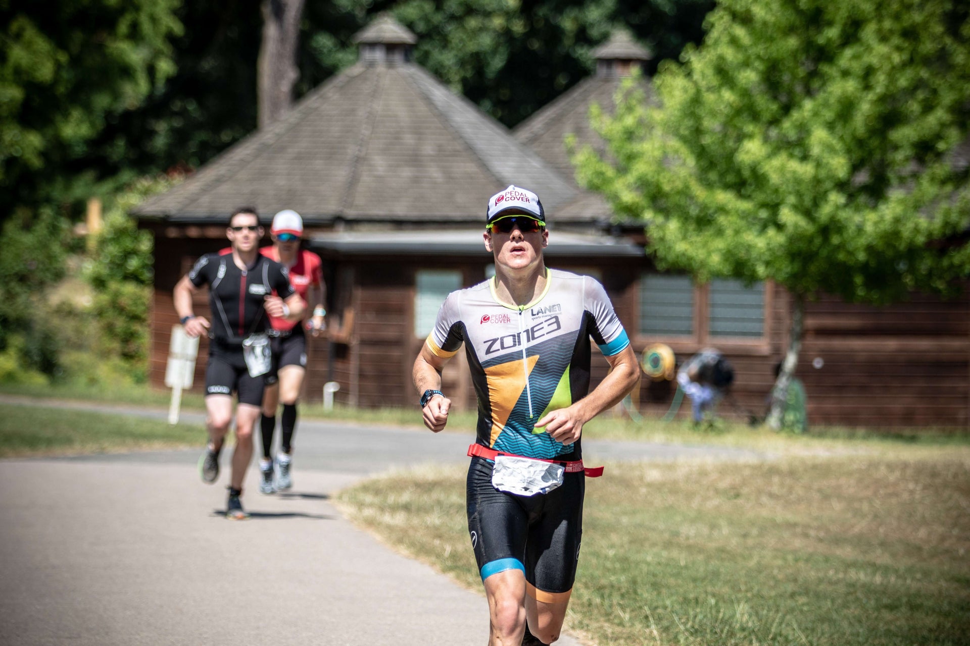 Front on triathlete in white and colorful kit runs along a country road with a wooden lodge and trees behind