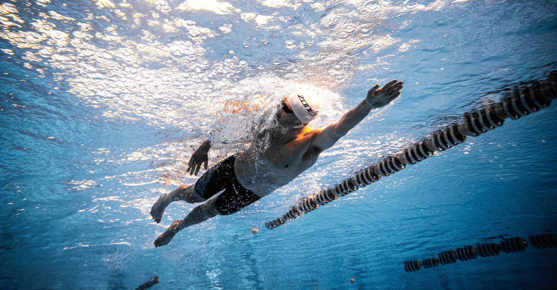 Swimmer in a pool wearing a white cap and dark wetsuit gliding along a lane.