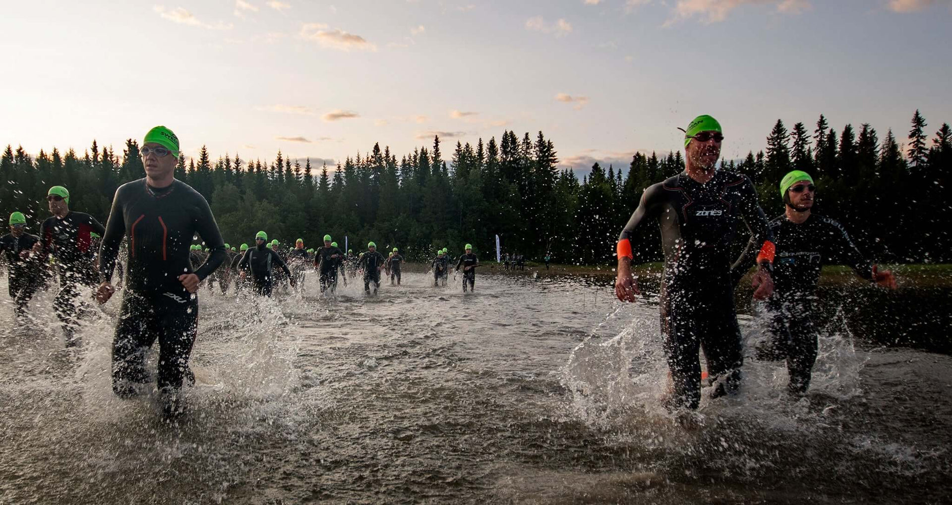 Group of athletes in wetsuits with bright green swim caps entering the water at a forested open water start.