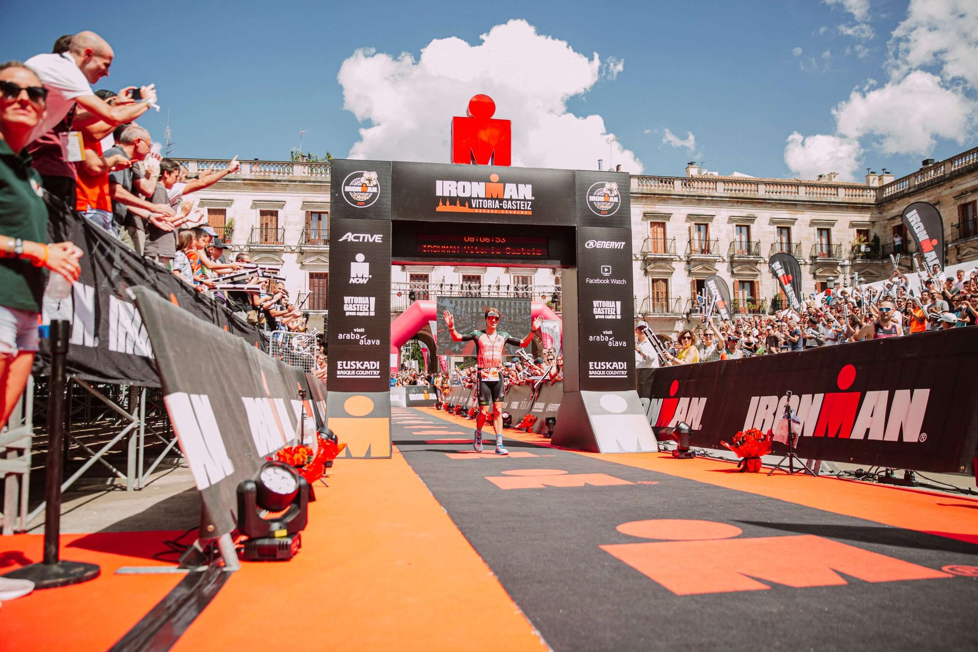 Triathlete crosses the Ironman finish line on an orange carpet as a cheering crowd fills a sunny city square