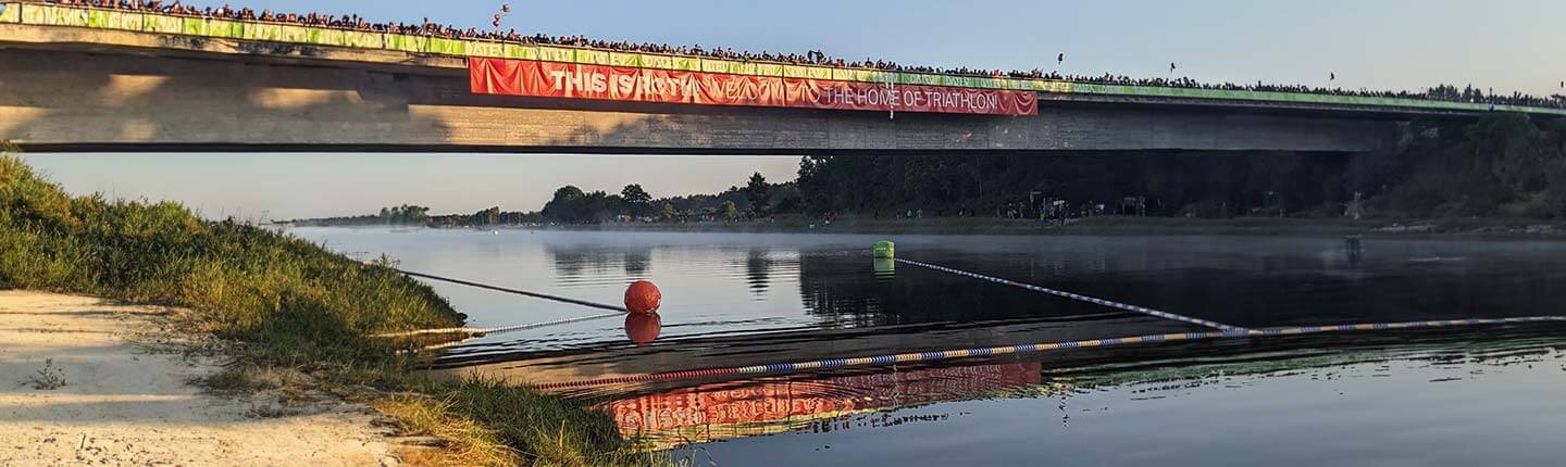 Spectators crowd a bridge over a calm river during a triathlon with a long red banner across the span