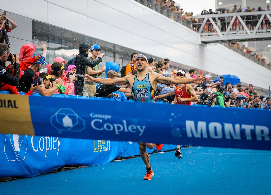 Runner in blue kit crosses the finish line with arms wide as fans and photographers cheer behind blue barriers.