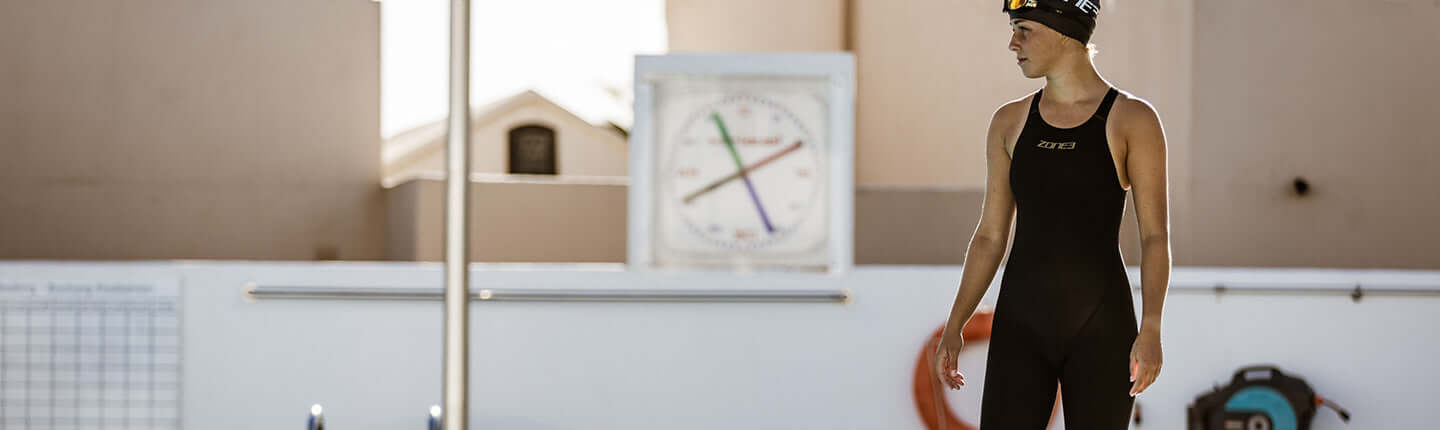 Swimmer in a black full body wetsuit and cap stands poolside near a large clock.