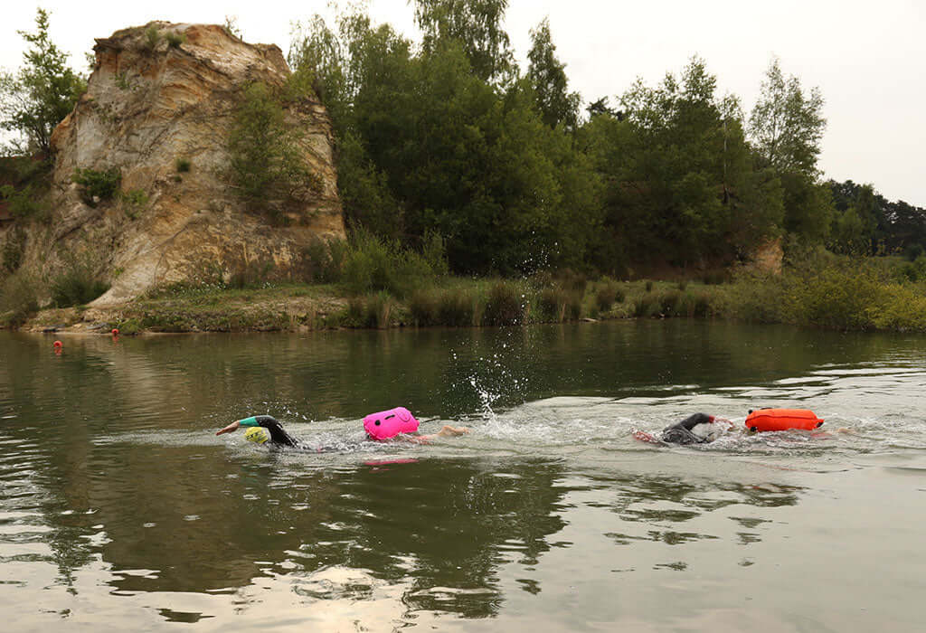 Open water swimmers with bright flotation devices glide across a calm river beside a rocky bank and trees.