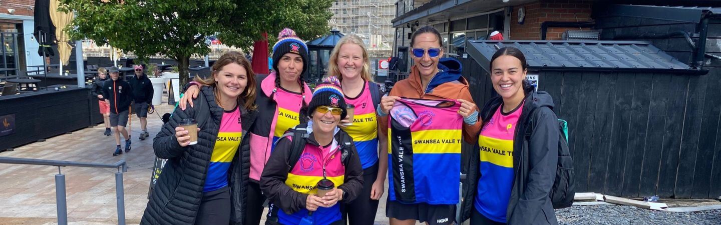 Swansea Vale Tri club members in bright jerseys pose together by a waterfront outdoor area.