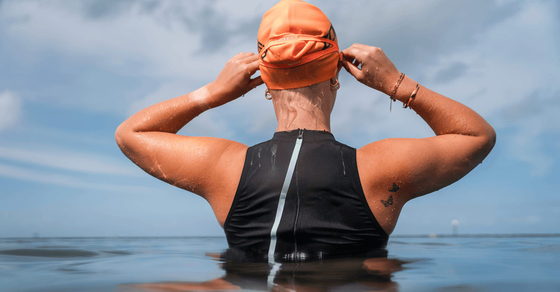 Back view of a swimmer in a black and white triathlon suit adjusting an orange swim cap outdoors.