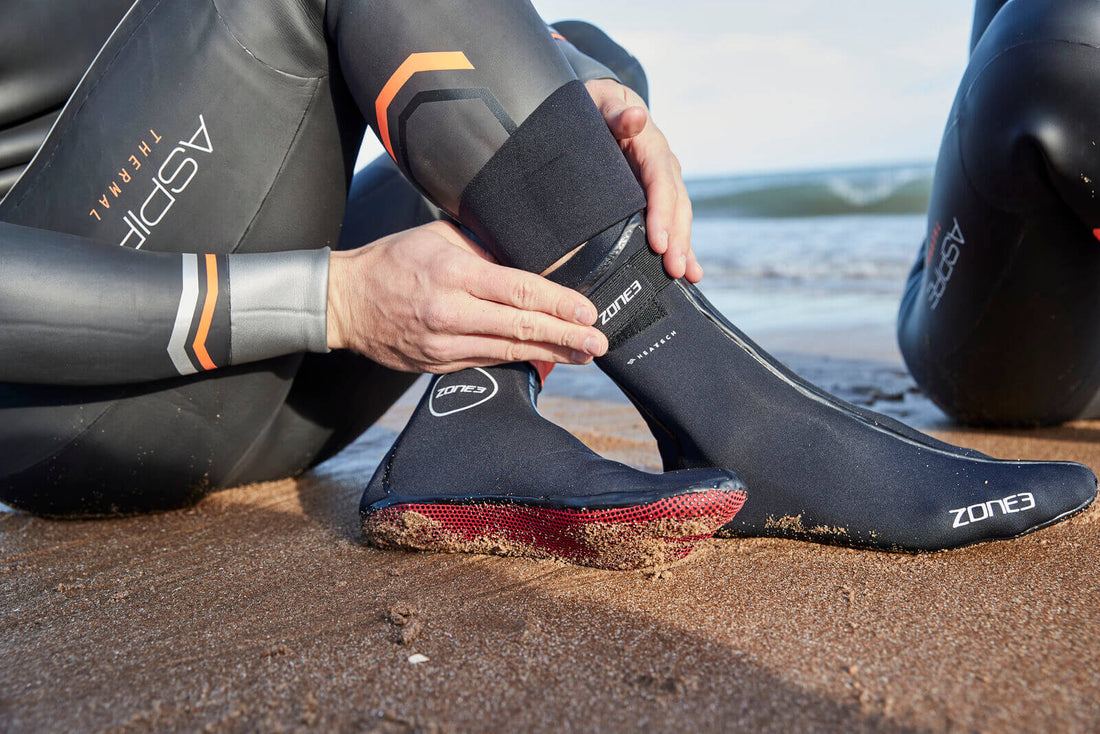 Person on a sandy beach adjusts Zone3 neoprene swim socks with waves in the background.
