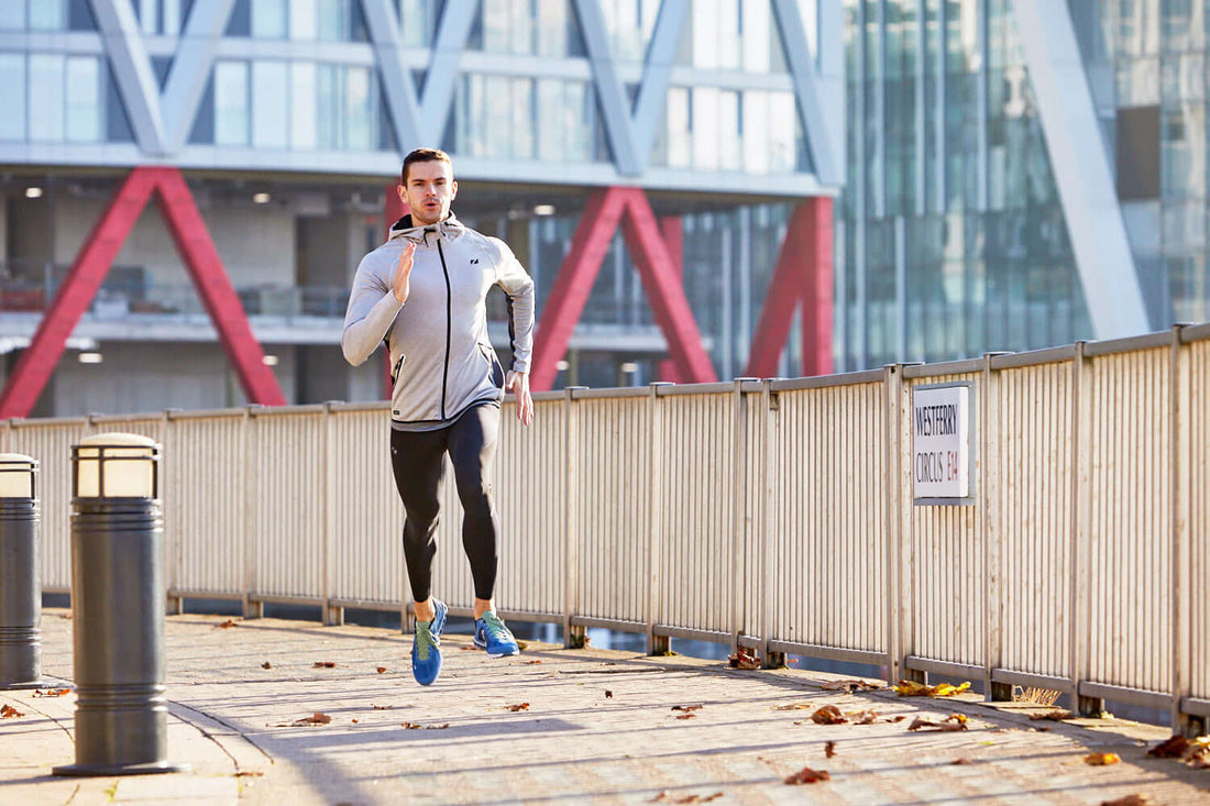 Male runner in a gray jacket and blue shoes sprints along a waterfront promenade with modern glass buildings in the background