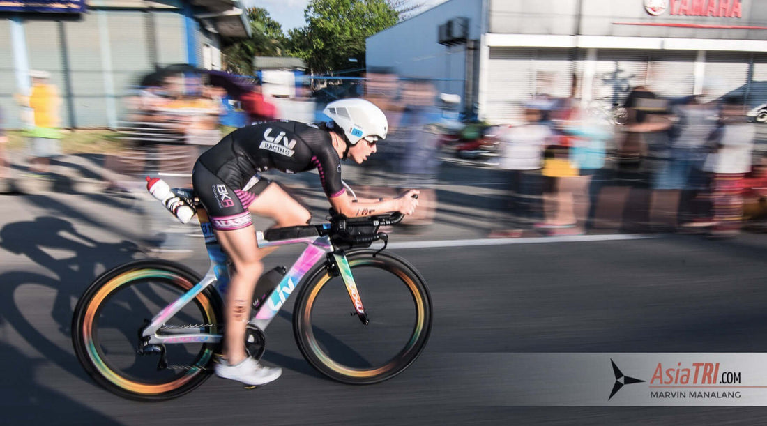 Female triathlete in black kit on a pastel gradient aero bike speeds past cheering spectators during IM 70.3 Davao.
