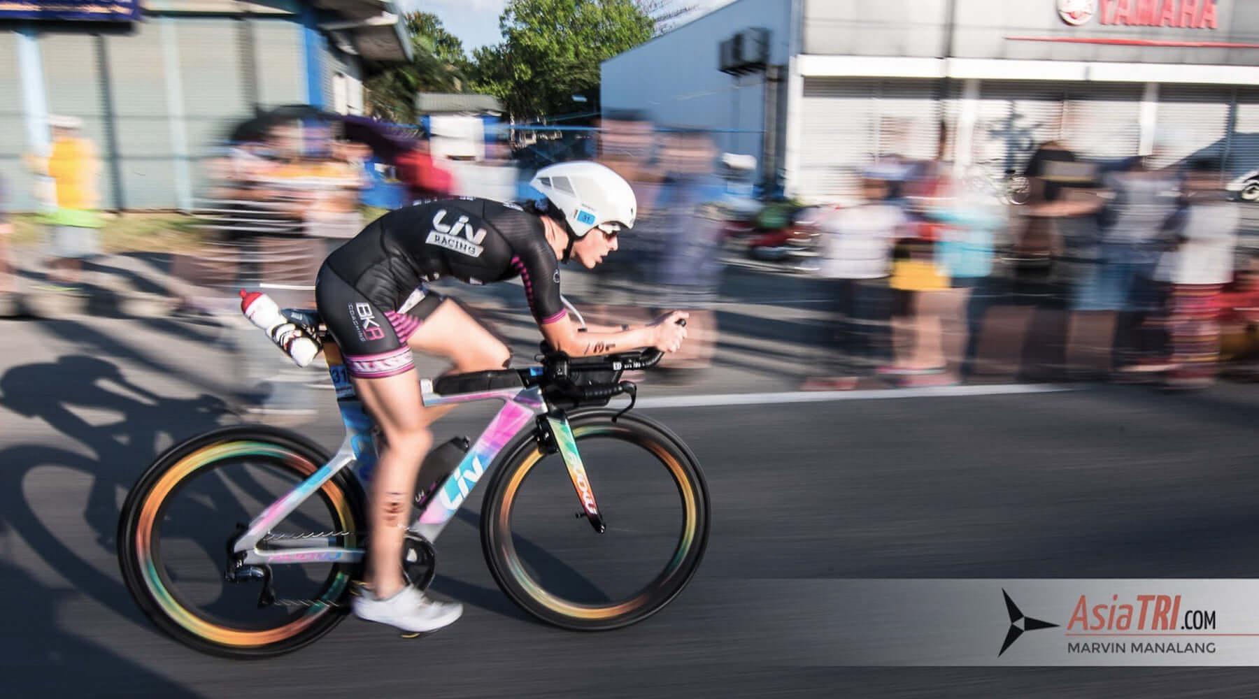 Female triathlete in black kit on a pastel gradient aero bike speeds past cheering spectators during IM 70.3 Davao.