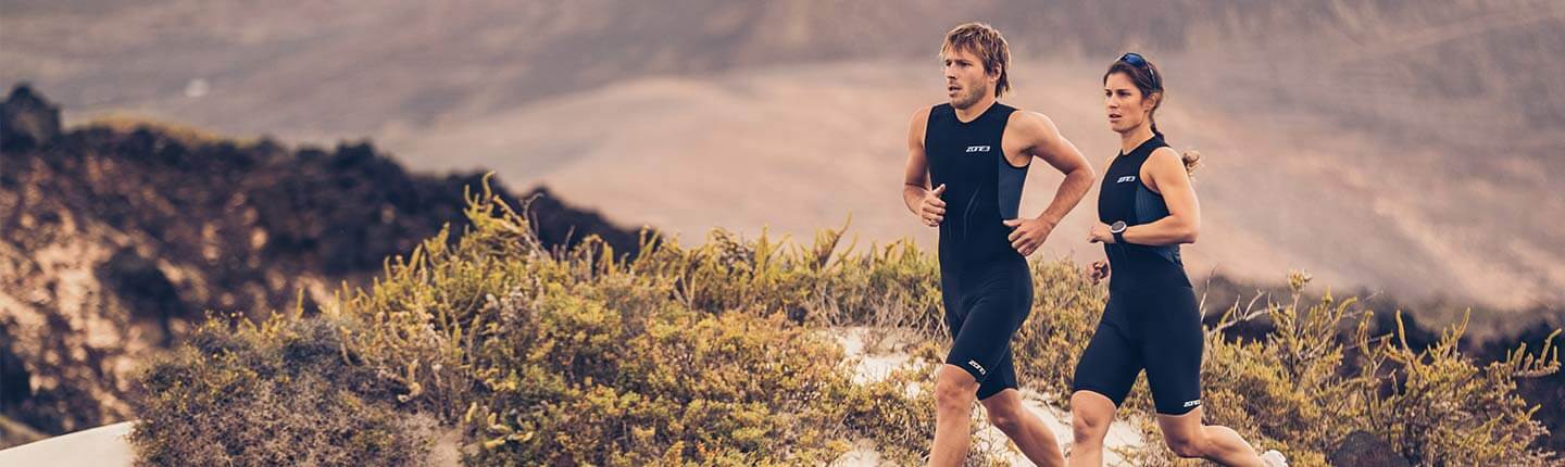Two athletes in matching black tri suits jog along a dry hillside trail