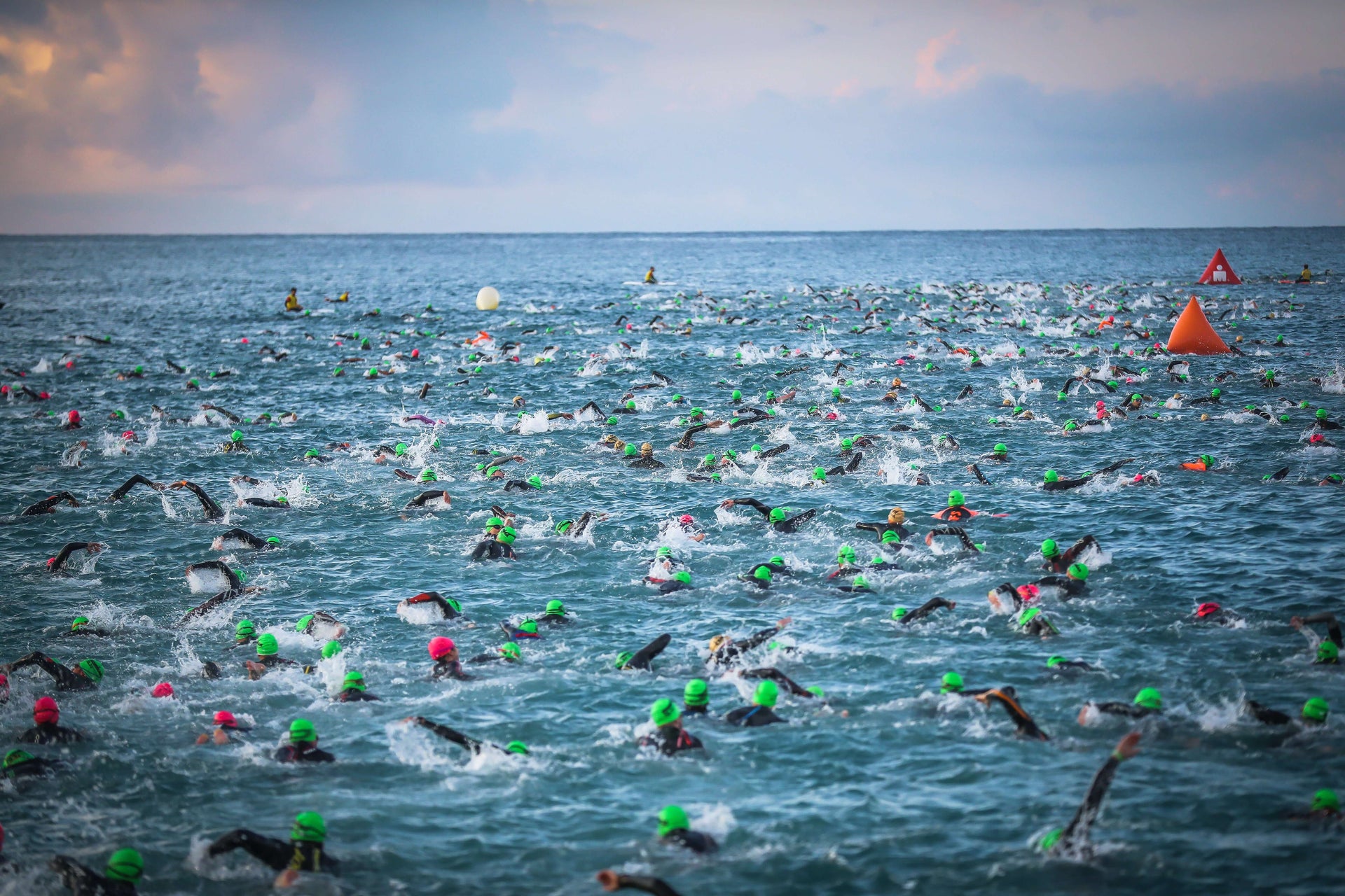 Hundreds of triathletes in green swim caps begin the open water swim as buoy markers float among the churning waves