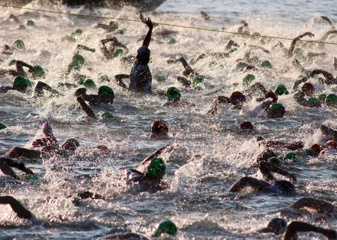 Open water triathlon start with swimmers in green caps; a marshal raises an arm above the waves beside an orange buoy.