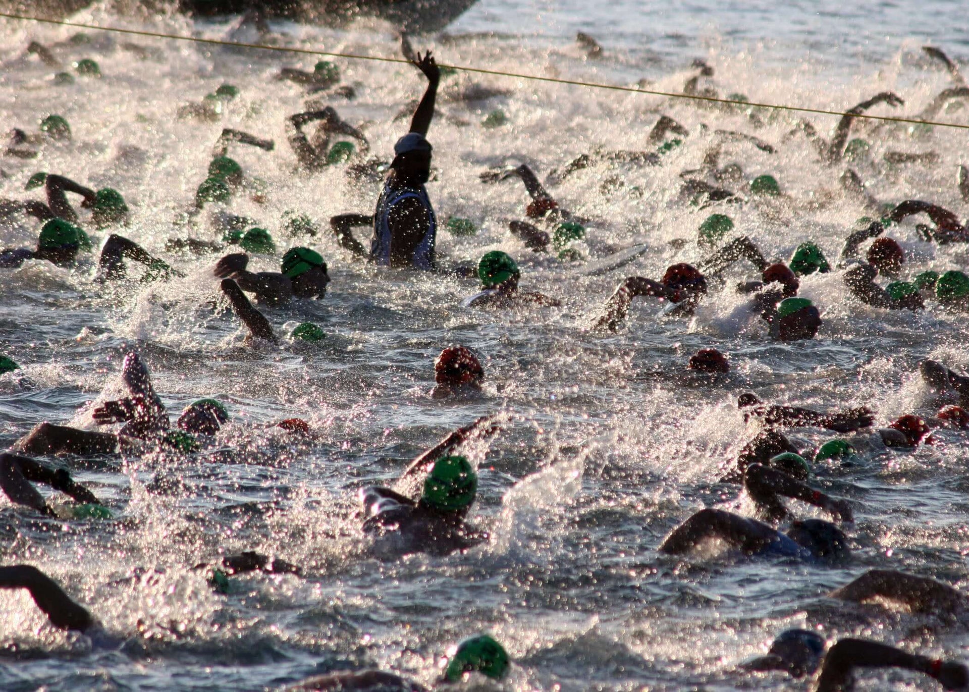 Open water triathlon start with swimmers in green caps; a marshal raises an arm above the waves beside an orange buoy.