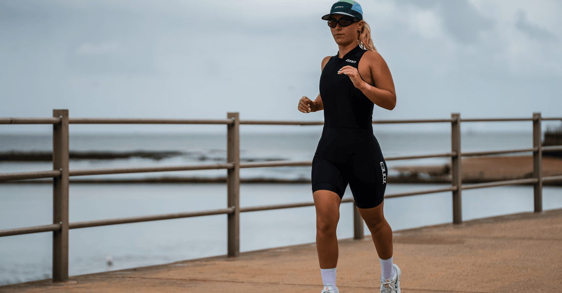 Female triathlete running along a seaside boardwalk in a black sleeveless trisuit with cap and sunglasses.