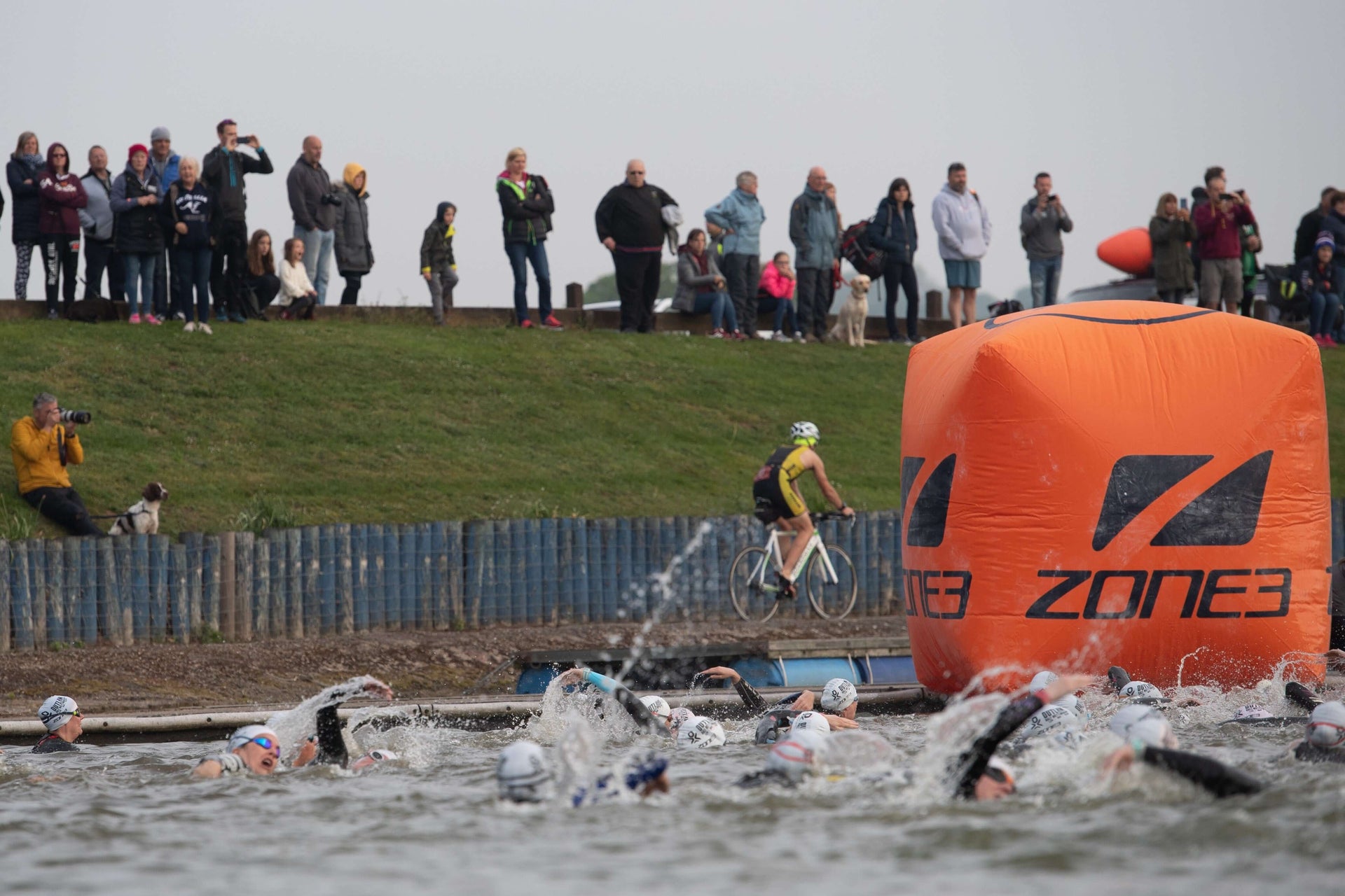 Spectators line a grassy embankment watching a race, with an orange inflatable barrier in the foreground.