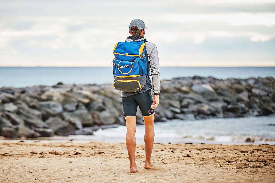 Back view of a person on a beach with a blue and yellow Zone3 kit bag looking toward the water