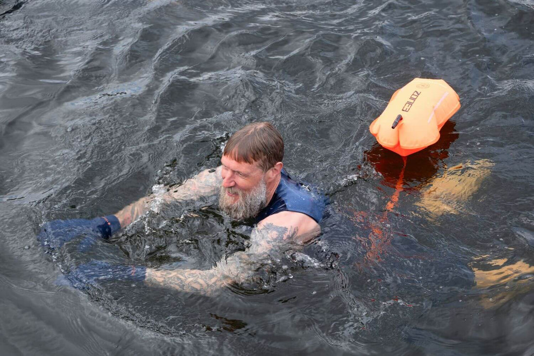 Bearded swimmer in a wetsuit top swims in open water with an orange safety buoy nearby.