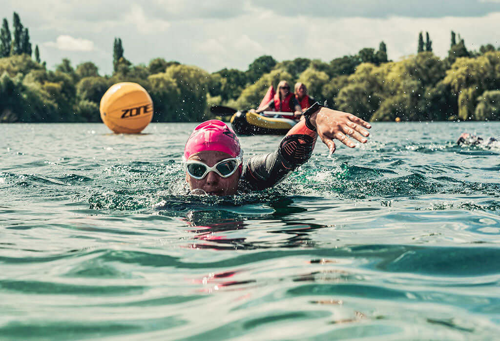 Swimmer with pink cap and goggles paddles in a lake with a yellow buoy and other swimmers in the distance.