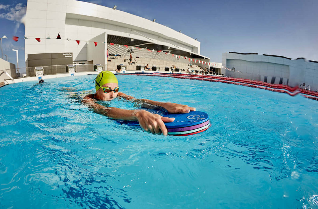 Swimmer wearing a lime green cap and goggles holds a blue circular flotation buoy in an outdoor pool