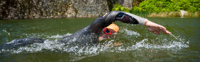 someone is swimming in a lake, breathing to one side whilst doing frontcrawl they are wearing the aspire wetsuit and orange swim cap and orange goggles