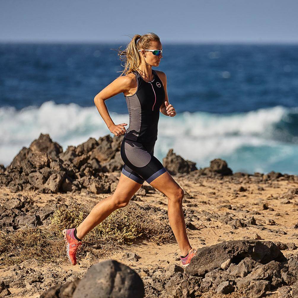 Athlete wearing a black gray women triathlon suit runs along rocky beach.