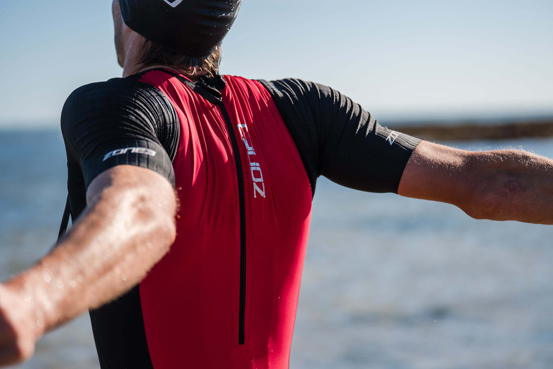 Athlete wearing a red and black short sleeve trisuit poses with arms raised by the beach.