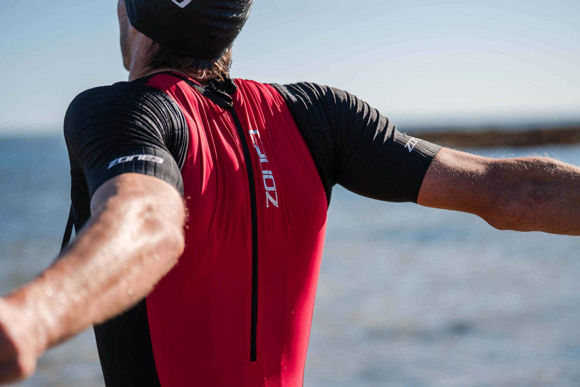 Athlete wearing a red and black short sleeve trisuit poses with arms raised by the beach.