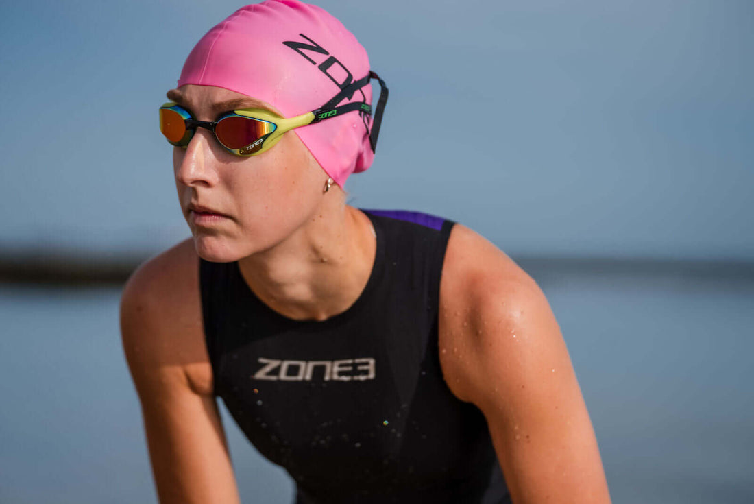 Swimmer wearing a pink swim cap and mirrored goggles stands by the water.