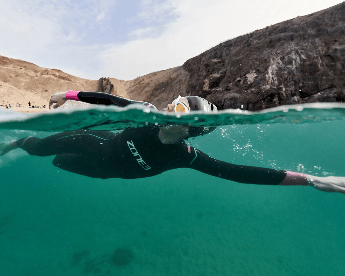 Half submerged shot of a swimmer in a black Zone3 wetsuit gliding through turquoise water near a rocky shore.