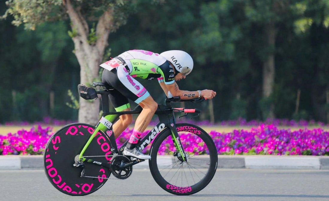 Triathlete in neon green and pink kit rides a time trial bike on a road with a flower bed in the background