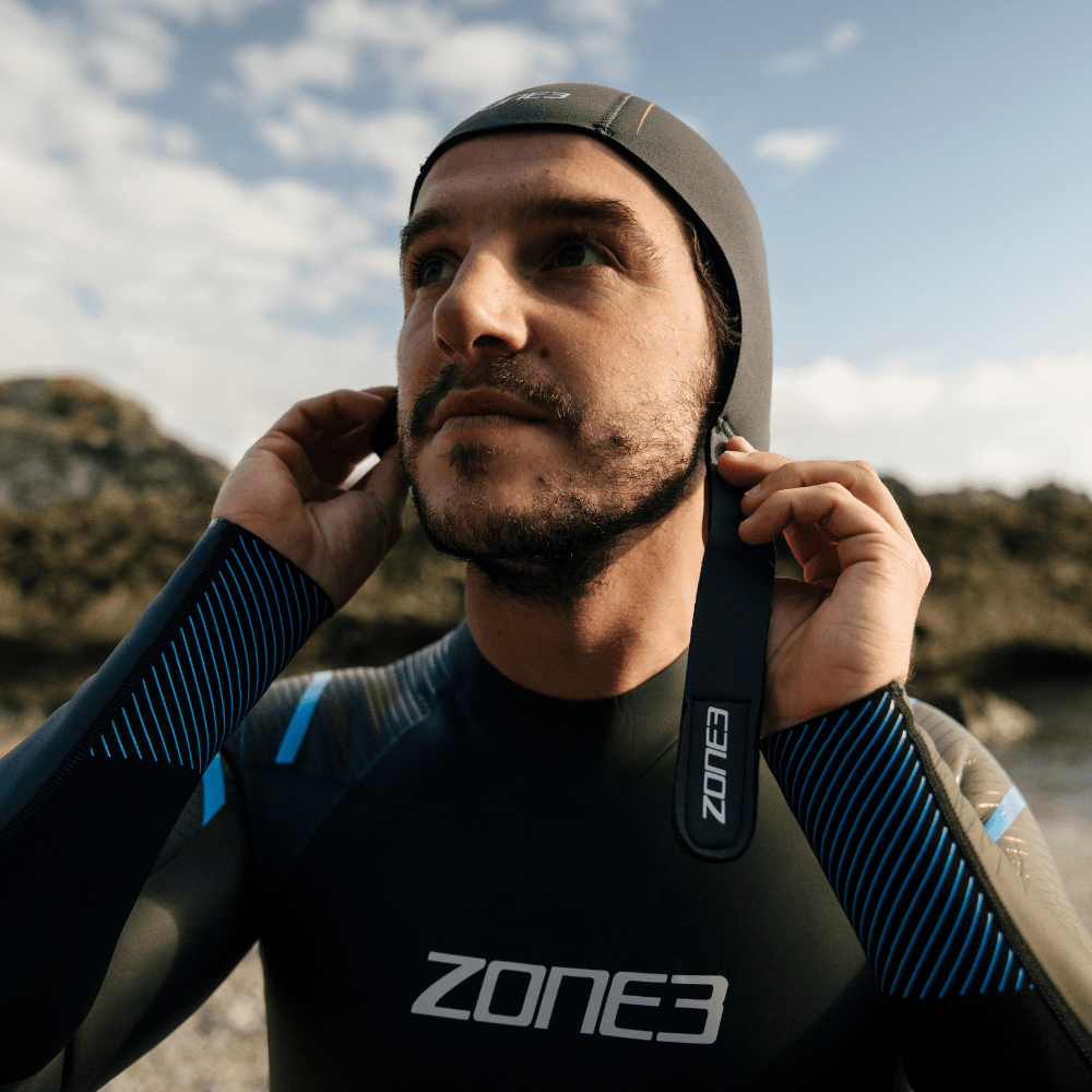 Man in a gray Zone3 neoprene thermal cap adjusts the strap of his wetsuit outdoors at the beach