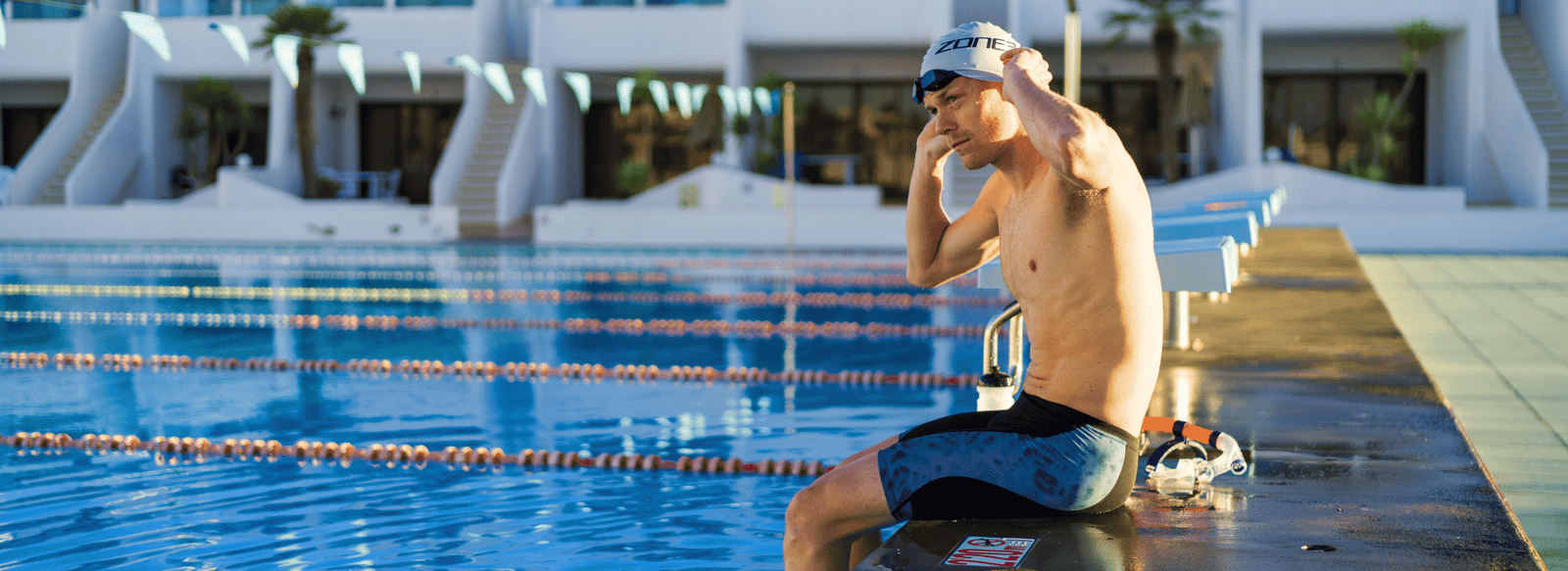 casper stornes sitting on the side of a swimming pool wearing jammers, adjusting swim cap