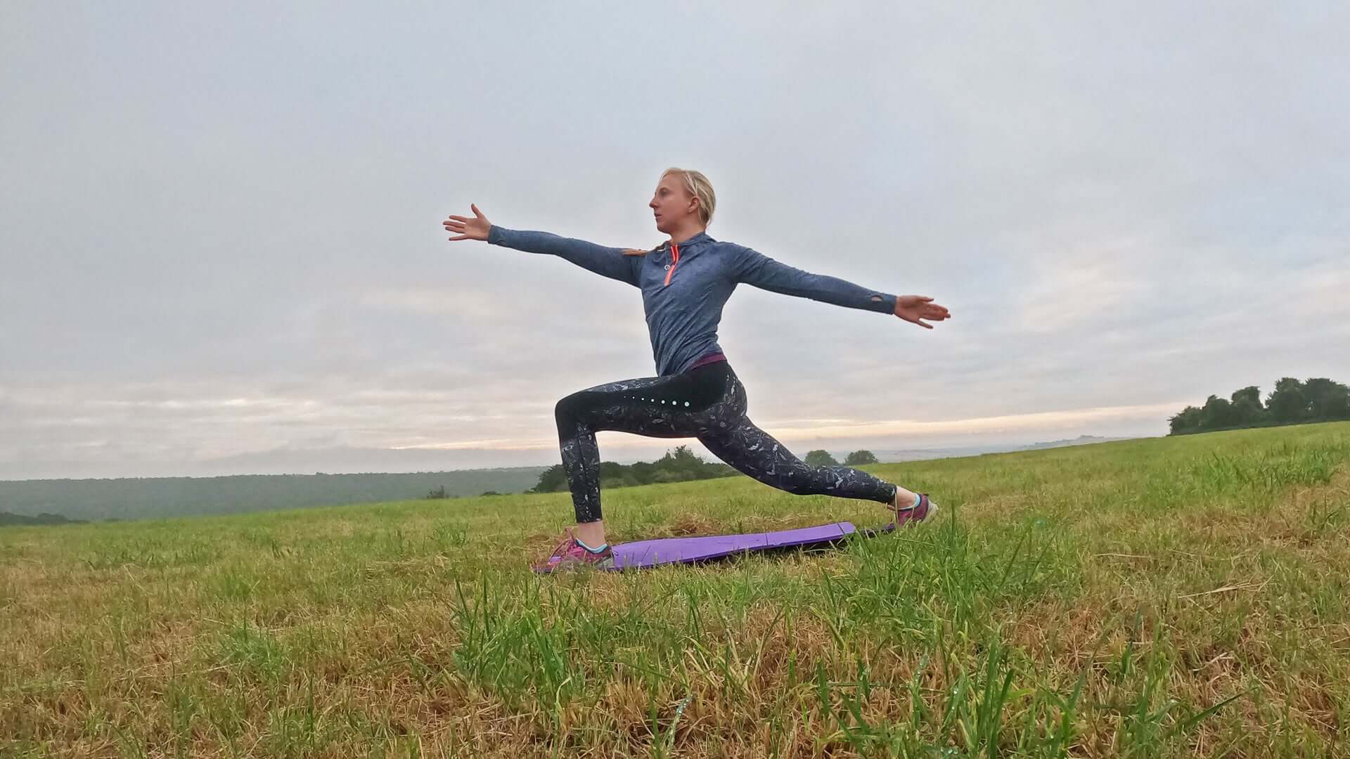 Woman in a blue long sleeve top and patterned leggings on a purple mat performing Pilates in a grassy field.