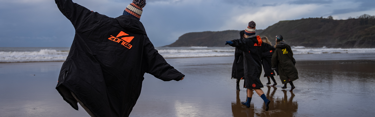 A group of swimmers are running on the sand at low tide towards the water - they are wearing zone3 changing robes to keep warm.