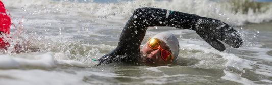 a woman is swimming in the sea wearing a wetsuit and zone3 neoprene gloves and orange polarised goggles