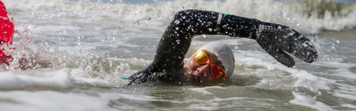a woman is swimming in the sea wearing a wetsuit and zone3 neoprene gloves and orange polarised goggles