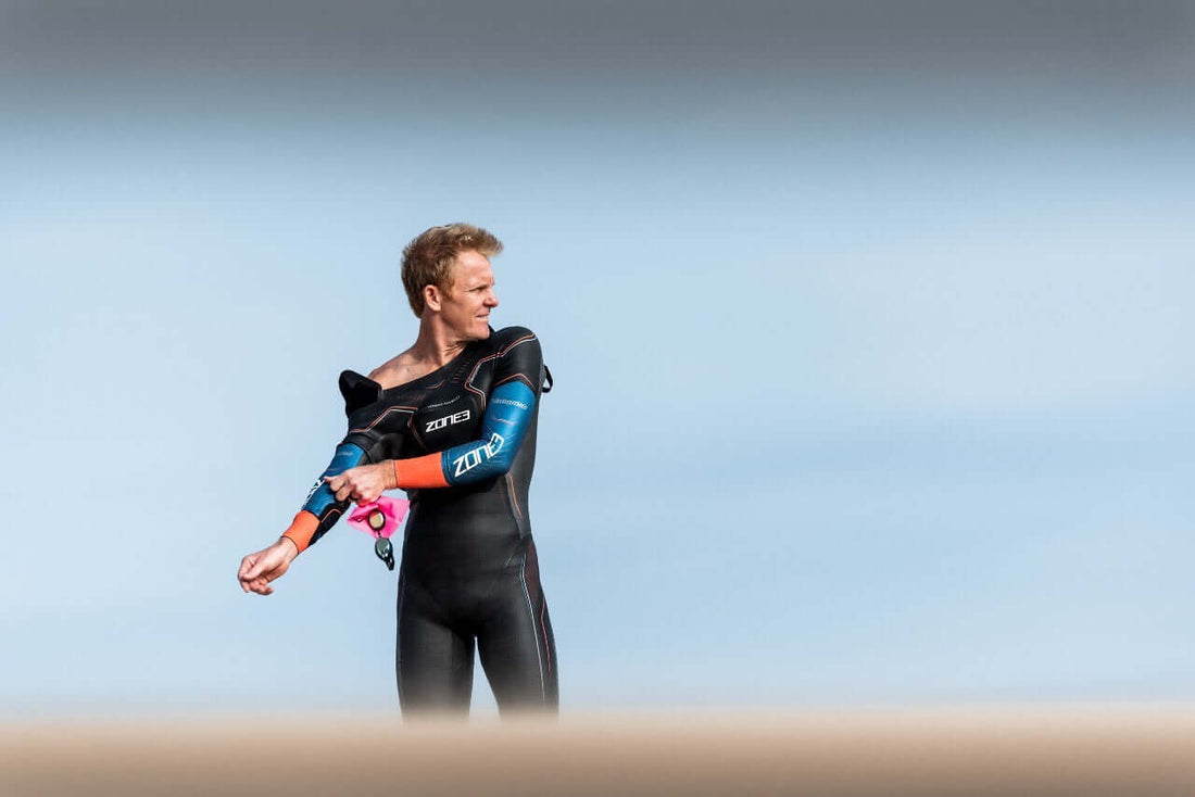 Triathlete in a Zone3 wetsuit with blue sleeves adjusts his sleeve against a pale blue sky
